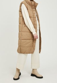Lichtgewicht, beige puffer bodywarmer met een hoge kraag, drukknopen en een doorgestikte textuur, gedragen over een crèmekleurig shirt met lange mouwen en witte broek.