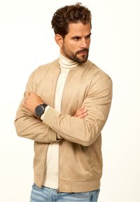 Tan suede bomber jacket with a ribbed collar and cuffs, zip closure, and side pockets, worn over a cream turtleneck sweater.