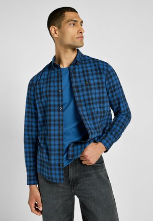 Camisa de cuadros azul y negra con botones y cuello, usada sobre una camiseta azul. La camisa tiene mangas largas y un logo en el pecho.