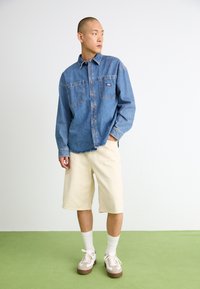 Lyseblå denimskjorte med to brystlommer, beige knelange shorts og brune og hvite joggesko. Gressgrønn gulvbakgrunn.