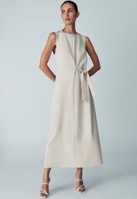 A long, sleeveless dress in light beige fabric with a soft drape. It features a front seam and a tied waist detail. Simple and elegant design.