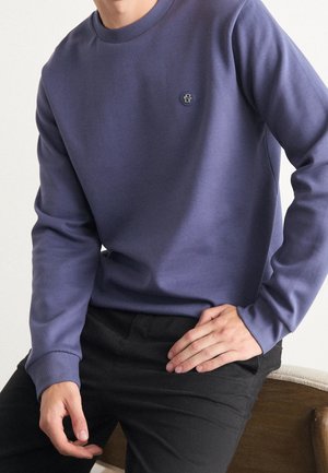 Sweatshirt long violet à manches longues avec un col rond, featuring un petit logo brodé sur la poitrine. Porté avec un pantalon noir.