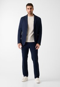 Marineblauw kostuumjasje en broek met beige t-shirt. Gladde stof, getailleerde pasvorm, twee voorzakken en witte sneakers. Eenvoudig en modern ontwerp.