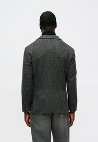 Hallhalli vääristatud villane bleiser, millel on kahe nupuga kinnitamine ja lõikega kraed, koos heleda halli teksaga. Kohandatud lõige, tekstureeritud viimistlus.