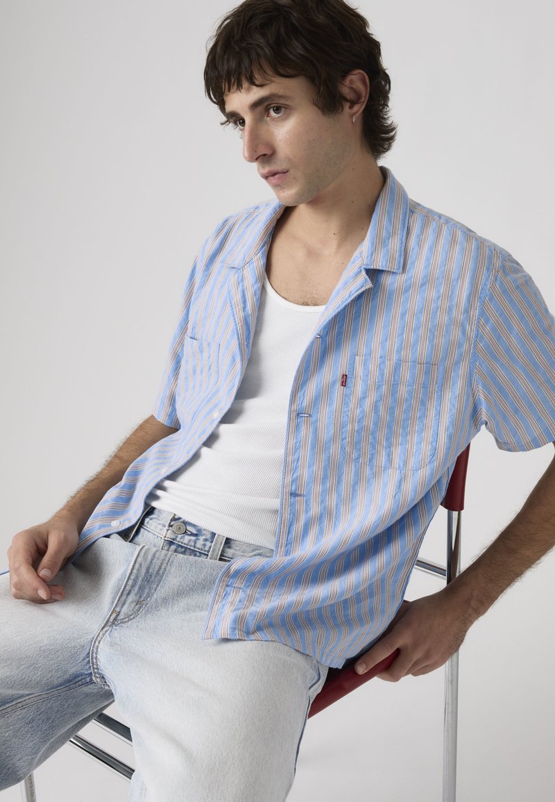 Joven con cabello oscuro que lleva una camisa de manga corta a rayas azules, camiseta blanca sin mangas y vaqueros de lavado claro sentado en una silla roja.