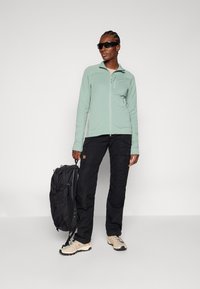 Hellgrüne Zip-Jacke, schwarze Cargo-Hosen, beige Wanderschuhe und ein schwarzer Rucksack. Glatte Stofftextur, praktisches Design mit Taschen.
