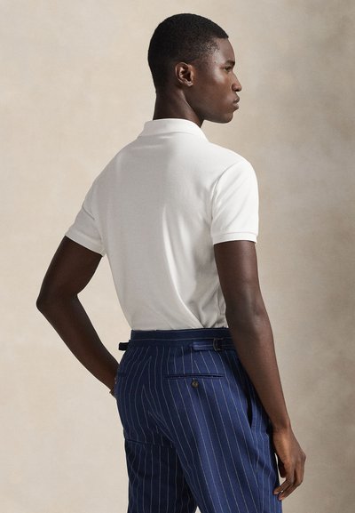 Mand iført en hvid kortærmet poloshirt og blå bukser med nålestriber, stående i profil mod en neutral baggrund.