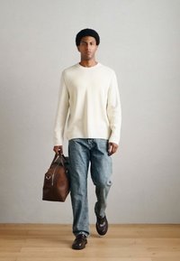 Bēšs adīts džemperis, zilas džinsa bikses, brūna ādas soma. Modelis valkā tumšas kurpes bez šņoru, stāvot uz gaiša koka grīdas ar neitrālu fonu.