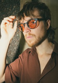 Γυαλιά ηλίου με μαύρο σκελετό και πορτοκαλί φακούς, που φοριούνται από ένα άτομο με κοντά μαλλιά και γένια, με φόντο υφή.