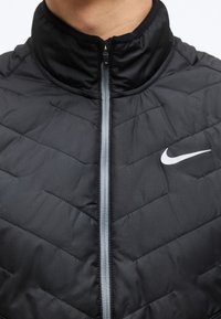 Μαύρο καπιτονέ μπουφάν με εμπρόσθιο φερμουάρ και λευκό λογότυπο Nike swoosh στο αριστερό στήθος, που φοριέται από άτομο.