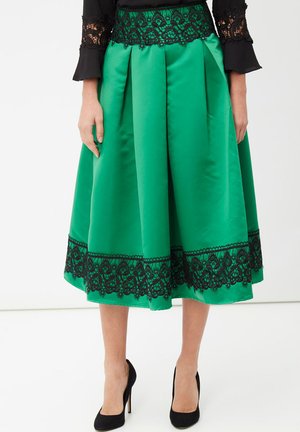 Falda midi de satén verde con ribete de encaje negro en la cintura y el dobladillo, combinada con tacones altos negros y un top negro con puños de encaje.