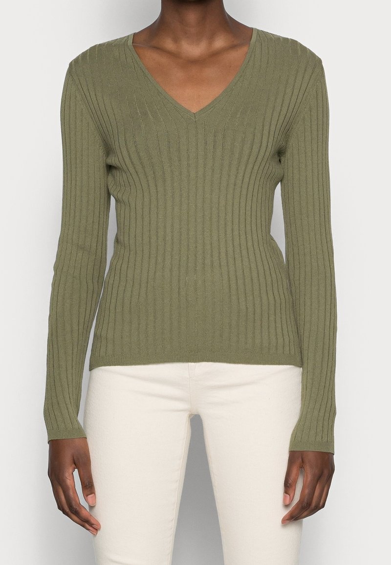 Personne portant un pull ajusté côtelé à manches longues col V de couleur vert olive et un pantalon blanc, debout les bras détendus le long du corps.