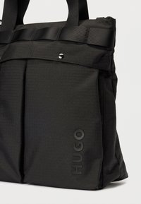 Svart teksturert stoff tote bag med doble håndtak, frontlomme med trykknapp, og "HUGO" merke navn preget vertikalt i hjørnet.