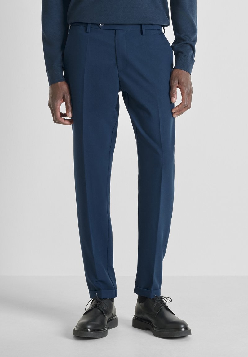 Muškarac nosi tamnoplave krojene hlače s manžetama i crne kožne cipele na vezanje na jednostavnoj pozadini.