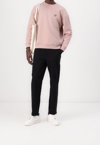 Man in een roze sweatshirt met een geel vogellogo, zwarte broek, witte sneakers en een beige draagtas over zijn schouder.