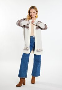 Lange Strickjacke in Hellgrau mit einem geometrischen Muster in Braun und Creme. Kombiniert mit einem hellbeigen Oberteil und ausgestellten Jeans in Denim. Braune Stiefeletten.