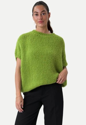 Woman wearing a loose-fitting, bright green knitted short-sleeve sweater and black pants, standing against a plain background.