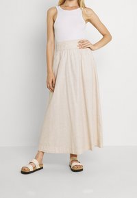 Jupe maxi en tissu beige clair avec une texture à motif subtil, dotée d'une taille élastique. Associée à des sandales blanches avec une semelle en liège.