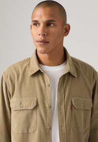 Tan corduroy shirt met twee voorzakken, knoopsluiting en een rood logo label. Draagt over een witte ronde hals t-shirt.