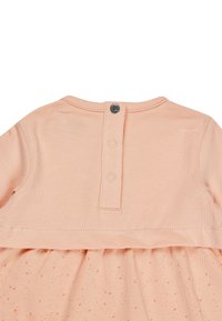 Pfirsichfarbenes Kleid mit kurzen Ärmeln, aus weichem Baumwollstoff, mit gerafftem Saum und dreifacher Knopfverschluss hinten. Subtiles Punktemuster nahe dem Saum.