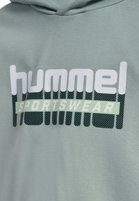 Sweat à capuche en tissu vert clair. Présente un grand graphisme blanc et vert "hummel SPORTSWEAR" avec des points texturés et des lignes horizontales.