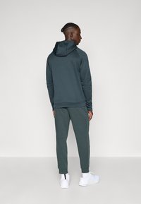 Donkergroen hoodie en bijpassende joggers van zachte stof. De hoodie heeft een voorzak en ribbels aan de mouwen, gecombineerd met witte sneakers.