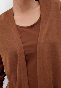 Hnědý cardigan s otevřeným předním dílem, nošený přes tričko se stejným odstínem a kulatým výstřihem. Měkká textura s jemným vzorem pletení.