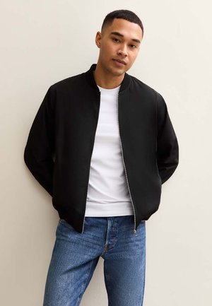 Jeune homme portant une veste noire à fermeture éclair, un t-shirt blanc et un jean bleu, debout avec les mains derrière le dos contre un mur uni.