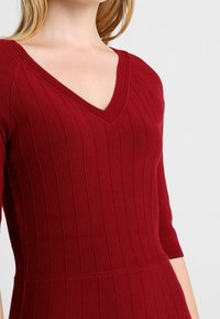Abito ribbed color bordeaux con scollo a V, maniche a tre quarti e vita aderente. Il materiale appare morbido ed elastico con righe verticali.