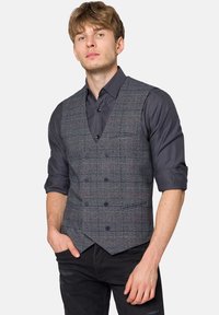 Szary kamizelka w kratę z czerwonymi akcentami, z dekoltem w serek i pięcioma guzikami, noszona na ciemnej koszuli; stylizowana z czarnymi dżinsami.