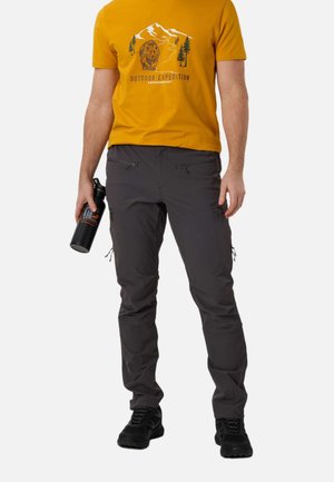 Mand i sennepsgul "Outdoor Expedition" t-shirt, mørkegrå vandrebukser med lynlås, sorte sko, der holder en sort vandflaske.