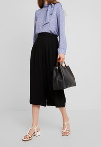 Kvinna som bär en blå mönstrad blus och svarta culotter, håller en svart handväska med sköldpaddsmönstrade handtag, bär vita klacksandaler.