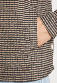 Main dans la poche d'une veste à motif pied-de-poule marron, vert et beige avec une petite étiquette blanche, sur un jean en denim bleu clair.
