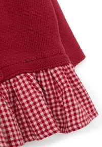 Haut en tricot rouge avec un ourlet à volants. L'ourlet présente un motif vichy rouge et blanc. Texture lisse sur le haut, texturée au niveau du volange.