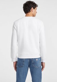 Hvit crewneck sweatshirt med ribbestrikkede mansjetter og kant, vist fra baksiden, kombinert med lyseblå jeans med et triangel-logo på lommen.