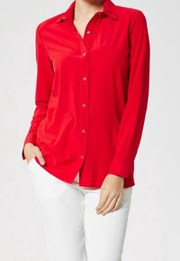 Chemise rouge à manches longues en tissu lisse, avec fermeture à boutons sur le devant, col pointu, et coupe ajustée, portée avec un pantalon blanc.