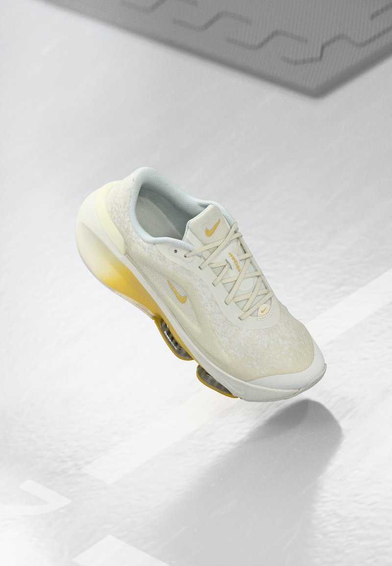 Witte en gele sportschoen met een gestructureerde bovenkant, vetersluiting en zichtbare luchtkussens. Gouden Nike-logo op de zijkant.