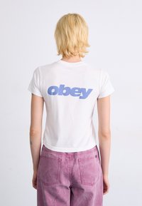 Balta trumpa rankove marškinėliai su šviesiai mėlynu užrašu "obey" ant nugaros, derinami su rožinėmis, aukštu liemeniu kelnėmis. Lygi audinio tekstūra.