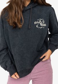Žena v bledě černé mikině Billabong a růžových kordových kalhotách, s dlouhými hnědými vlasy, drží okraj mikiny v rukou.