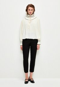 Weißes Blusenshirt mit Falten, hochgeschlossen und mit Ripsband; schwarze schmale Hose; schmaler Schnitt; glatte Textur; schwarze Schuhe mit Knöchelriemchen.