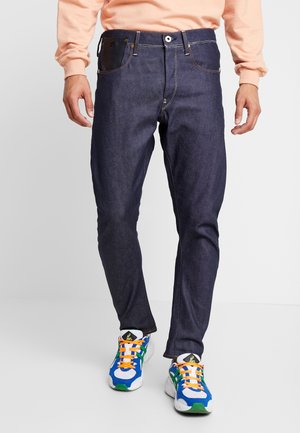 Muž nosící tmavě modré džíny, broskvovou mikinu a barevné modré, zelené, bílé tenisky s oranžovými tkaničkami, stojí na jednobarevném bílém pozadí.