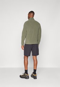 Grøn fleecejakke med høj krave, parret med sorte shorts. Model iført kraftige vandresko med orange detaljer. Enkle baggrund.