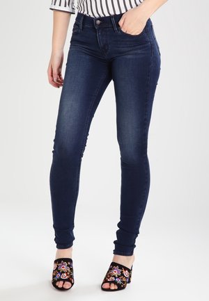 Smaltsiddende mørkeblå jeans båret med sorte åbne hæle med farverig blomsterbroderi. Én hånd hviler i forlommen.