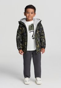 Wyściełana kurtka w ciemnozielonym kolorze z wzorem dinozaurów, szara kaptura, biała koszulka z grafiką dinozaura, szare spodnie i białe sneakersy.