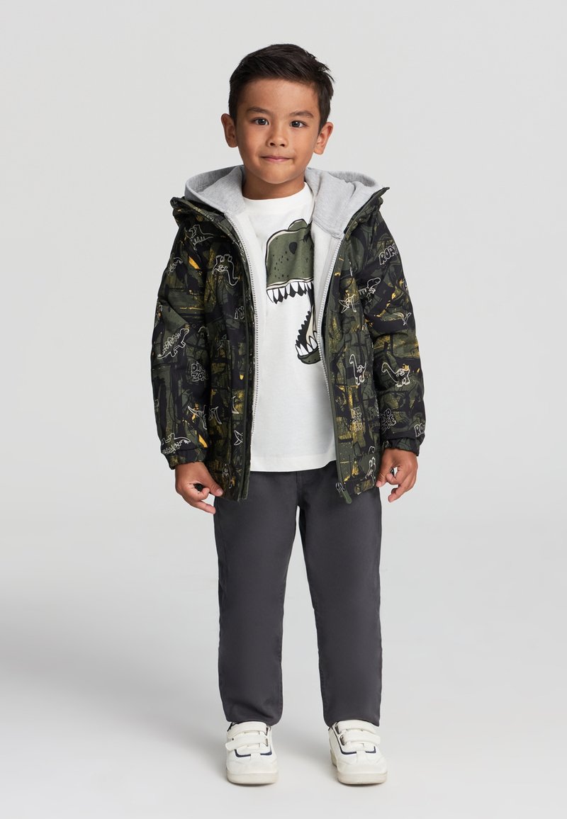 Wyściełana kurtka w ciemnozielonym kolorze z wzorem dinozaurów, szara kaptura, biała koszulka z grafiką dinozaura, szare spodnie i białe sneakersy.