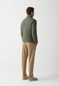 Kevyt vihreä untuvatakki vaakasuoralla tikkauskuvalla, korkea kaulus ja sivutaskut, yhdistettynä beigeihin kapeneviin housuihin ja monivärisiin tennareihin.