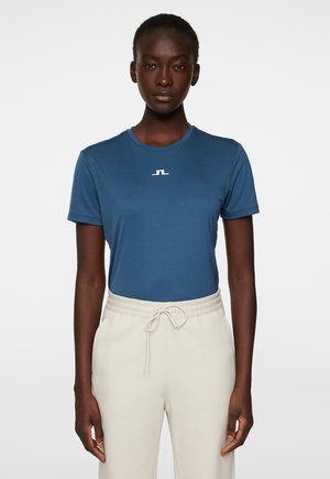 T-shirt blu a maniche corte in tessuto morbido, con un piccolo logo bianco sul petto, abbinato a pantaloni chiari in beige con coulisse.