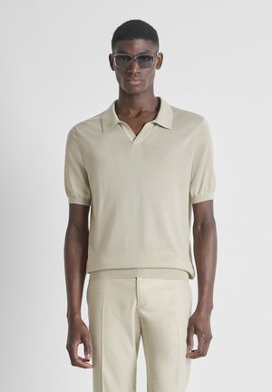 Hombre joven con camisa tipo polo de manga corta beige, pantalones a juego y gafas de sol oscuras, de frente contra un fondo liso.