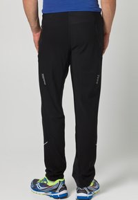 Mann in schwarzen winddichten Sporthosen und bunten Laufschuhen, mit dem Rücken zur Kamera stehend.