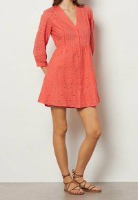Robe corail avec un décolleté en V, des manches trois-quarts, des détails en œillets brodés, une taille cintrée et une jupe évasée. Associée à des sandales à lanières marron.